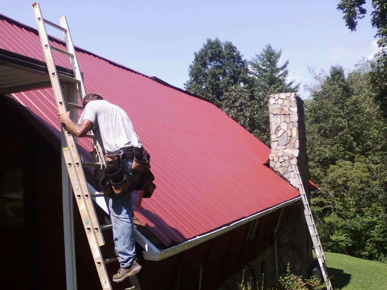 Roofer on a ladder beside a red metal roof and stone chimney — Soffit & Fascia Repair by Roofing Brothers in New Canaan